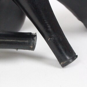 May include: Close-up of black high-heeled shoes. The image focuses on the heels, showing their shape and the wear and tear on the bottom. The shoes are on a white surface. The heels are thick and sturdy.