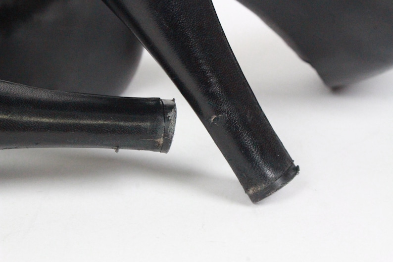 May include: Close-up of black high-heeled shoes. The image focuses on the heels, showing their shape and the wear and tear on the bottom. The shoes are on a white surface. The heels are thick and sturdy.