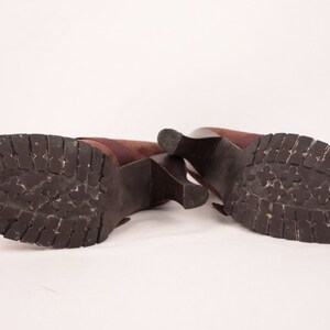 May include: A pair of brown heeled boots with a dark brown, textured sole. The boots have a block heel and a strap detail. The soles show wear and tear, suggesting they have been used.