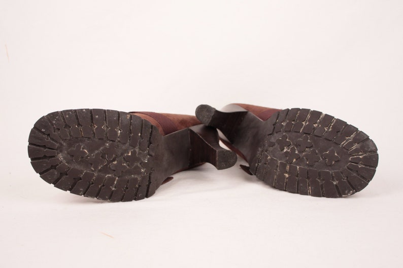 May include: A pair of brown heeled boots with a dark brown, textured sole. The boots have a block heel and a strap detail. The soles show wear and tear, suggesting they have been used.