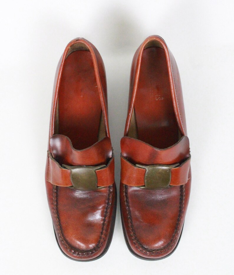 May include: A pair of brown leather loafers with a buckle detail. The loafers have a pointed toe and a low heel.