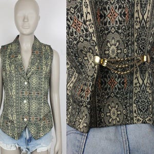 Puede incluir: Un chaleco vintage con un diseño estampado en tonos verdes, grises y melocotón. El chaleco tiene un cuello con solapas, cierre de botones y un cierre dorado decorativo con detalle de cadena en la espalda. El chaleco se exhibe en un maniquí.