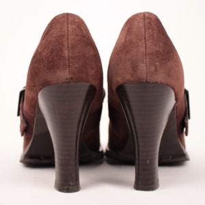 May include: A pair of brown suede high-heeled shoes. The shoes have a rounded toe, a buckle closure, and a dark brown, curved heel. The heels are approximately 3 inches (7.6 cm) high.