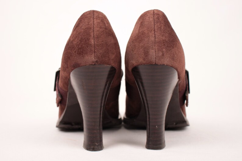 May include: A pair of brown suede high-heeled shoes. The shoes have a rounded toe, a buckle closure, and a dark brown, curved heel. The heels are approximately 3 inches (7.6 cm) high.