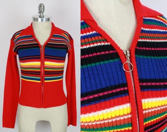 Op de afbeelding: Een vintage rode trui met ritssluiting en een kleurrijk gestreept ontwerp over de borst en bovenmouwen. De strepen zijn onder meer blauw, geel, zwart en wit. De trui heeft een kraag en lange mouwen.