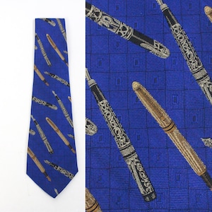 Puede incluir: Una corbata de seda azul con un patrón repetido de plumas estilográficas doradas y plateadas. Las plumas están dispuestas en diagonal a través de la corbata.