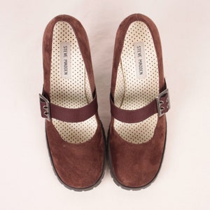 May include: A pair of brown suede Mary Jane style shoes with a strap and buckle closure. The interior features a white polka dot pattern and the brand name "STEVE MADDEN". The shoes have a chunky sole.