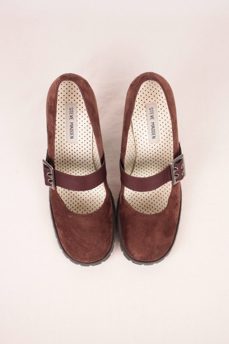 May include: A pair of brown suede Mary Jane style shoes with a strap and buckle closure. The interior features a white polka dot pattern and the brand name "STEVE MADDEN". The shoes have a chunky sole.