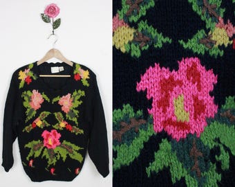 Op de afbeelding: Zwarte gebreide trui met een bloemenpatroon. De trui heeft roze, groene en gele bloemen die op de voorkant zijn geborduurd.
