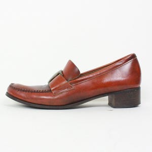 May include: Brown leather heeled loafers with a buckle detail. The loafers have a low block heel and a pointed toe.