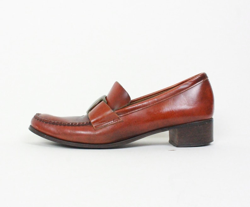 May include: Brown leather heeled loafers with a buckle detail. The loafers have a low block heel and a pointed toe.