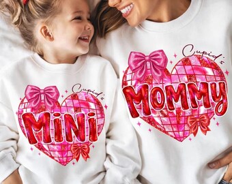 Pode incluir: Camisolas brancas com desenhos de coração a condizer. Uma diz "Mini" e a outra "Mommy" em vermelho, com laços cor-de-rosa e um padrão de grelha. As palavras "Cupid's" estão acima dos corações.