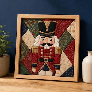Könnte beinhalten: Ein quadratisches Wandbild aus Stoff in einem Holzrahmen mit einem zentralen Nussknacker in einer rot-schwarz-goldenen Uniform. Der Hintergrund besteht aus einem geometrischen Patchwork aus gesteppten Stoffquadraten in festlichen Rot-, Grün-, Dunkelblau- und Cremetönen.