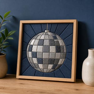Könnte beinhalten: Ein quadratisches Wandbild mit einem zentralen Discokugel-Motiv, das aus verschiedenen silbernen und grauen strukturierten Stoffquadraten besteht, vor einem dunkelblauen Stoffhintergrund mit strahlenden Linien, eingefasst in einen hellen Holzrahmen.