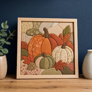 Könnte beinhalten: Ein quadratisches, gerahmtes Wandbild aus Stoff mit einem gesteppten Kürbismotiv. Das Kunstwerk zeigt drei Kürbisse in Orange, Creme und Salbeigrün, umgeben von gemusterten Stoffblättern in Rost-, Oliv- und Beigetönen, eingefasst in einen hellen Holzrahmen.