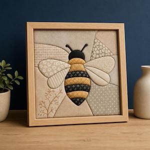 Könnte beinhalten: Ein quadratisches, gerahmtes Wandbild aus Stoff mit einem zentralen Bienenmotiv, das aus verschiedenen beigen, cremefarbenen und schwarzen gemusterten Textilstücken besteht. Die Biene hat einen schwarzen Kopf, Fühler und einen gestreiften Körper.