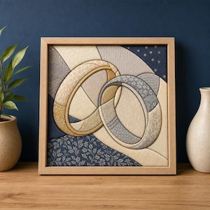 Op de afbeelding: Een vierkant ingelijst wandkunstwerk van stof met twee in elkaar grijpende ringvormen. Het ontwerp maakt gebruik van diverse gestructureerde stoffen in goud-, crème-, grijs- en marineblauwtinten, met verwerkte florale en geometrische patronen.