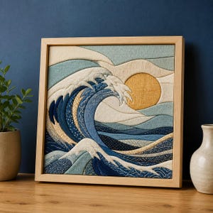 Könnte beinhalten: Ein quadratisches Wandbild aus Stoff in einem hellen Holzrahmen, das eine stilisierte Meereswelle und eine große gelbe Sonne zeigt. Das Kunstwerk verwendet geschichtete blaue, cremefarbene und gemusterte Stoffteile für eine strukturierte Optik.