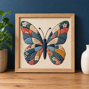 Op de afbeelding: Een vierkant ingelijst kunstwerk aan de muur met een vlinder gemaakt van stoffen lapjes in tinten blauw, rood, goud en crème. De vlindervleugels tonen diverse patronen, waaronder bloemmotieven en geometrische stippen op een beige stoffen achtergrond.