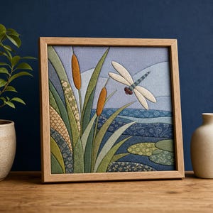 May include: A square fabric wall art piece in a light wood frame featuring a quilted scene of a dragonfly hovering over cattails and water. The composition uses various shades of green, blue, and tan fabric with floral and geometric patterns.