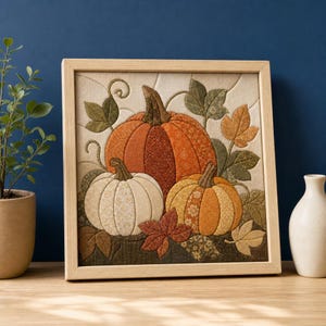 Puede incluir: Una pieza de arte mural de tela enmarcada de forma cuadrada con un diseño de aplique acolchado de tres calabazas en tonos naranja, crema y dorado. Las calabazas están rodeadas de hojas y enredaderas de tela en tonos verdes y marrones.