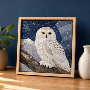 May include: A square fabric wall art piece in a light wood frame featuring a quilted snowy owl with yellow eyes perched on a branch. The background depicts a stylized mountain landscape and a dark blue night sky with white patterns and small dots.