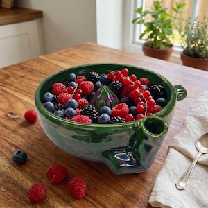 Könnte beinhalten: Eine grüne Zitruspresse aus Keramik, gefüllt mit frischen Himbeeren, Blaubeeren und Johannisbeeren. Die Schale verfügt über einen zentralen Presskegel und einen seitlichen Ausguss, mit glänzender Oberfläche und organischer Form.