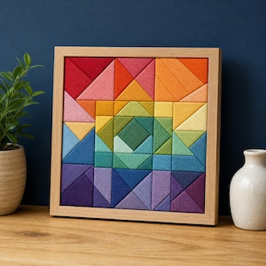 Könnte beinhalten: Ein quadratisches Wandbild mit einem geometrischen Mosaik aus Stoffdreiecken und -quadraten im Regenbogenspektrum, eingefasst in einen hellen Holzrahmen. Die Komposition zeigt ein zentrales Rautenmuster, das von verschiedenen Formen umgeben ist.