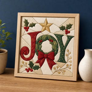 Op de afbeelding: Een vierkant ingelijst wandkunstwerk van stof met het woord JOY in een doorgestikte stijl. De letter J is rood, de O is een groene krans met een rode strik en de Y is groen. Een gouden ster bevindt zich boven de krans, met hulstbladeren en bessen.