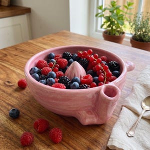 Könnte beinhalten: Eine rosafarbene Zitruspresse aus Keramik, gefüllt mit frischen Blaubeeren, Himbeeren und Johannisbeeren. Die Schale verfügt über einen zentralen Presskegel und einen seitlichen Ausguss. Lose Himbeeren und eine Blaubeere liegen auf dem Holz.