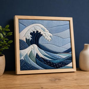 May include: A square textile wall art piece framed in light wood, featuring a fabric collage of a large ocean wave. The design uses various shades of blue and white fabric patches with different textures and patterns to depict the sea.