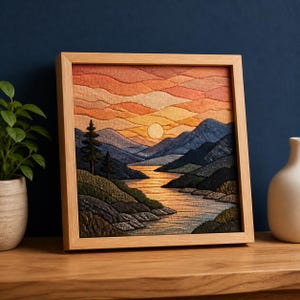 Puede incluir: Una pieza de arte textil cuadrada enmarcada en madera clara, que presenta un paisaje bordado de un atardecer sobre montañas y un río. La escena incluye nubes superpuestas en tonos naranja y amarillo, siluetas de montañas azul oscuro.
