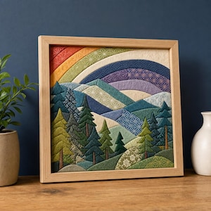 Könnte beinhalten: Ein quadratisches Wandbild aus Stoff in einem hellen Holzrahmen, das eine gesteppte Landschaft aus geschichteten Bergen und Kiefern zeigt. Die Szene umfasst einen farbenfrohen Regenbogenhimmel mit geometrischen Mustern in roten, gelben, blauen und violetten Stoffsegmenten.