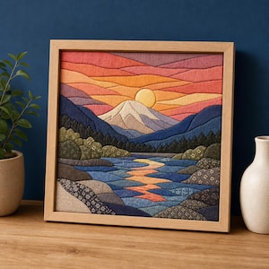 Puede incluir: Una pieza de arte mural de tela cuadrada en un marco de madera clara que representa un paisaje con una montaña, un sol poniente y un río. La escena utiliza parches de tela superpuestos en tonos naranja, rojo, azul y verde con patrones florales.