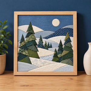 May include: A square fabric wall art piece in a light wood frame featuring a quilted winter landscape. The scene depicts layered snowy hills in shades of white and blue with various green patterned fabric pine trees and a pale moon.