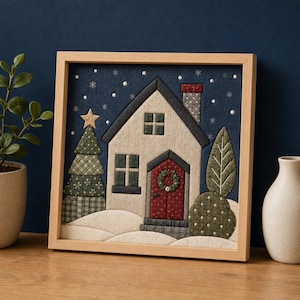 Könnte beinhalten: Ein quadratisches Wandbild aus Stoff in einem hellen Holzrahmen mit einer gesteppten Hausszene. Das Design zeigt ein cremefarbenes Haus mit roter Tür, einen grünen Weihnachtsbaum mit goldenem Stern und eine Schneelandschaft vor dunkelblauem Himmel.
