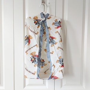 May include: A white garment bag with a repeating pattern of teddy bears wearing red and blue outfits. The bag has a blue denim-like trim and is hanging on a white hanger.