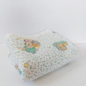 May include: A white baby blanket featuring a repeating pattern of green speckles, yellow stars, and cartoon characters including a mouse sitting on a crescent moon and a bear figure. The blanket has a white ruffled edge trim along the top border.