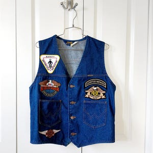 May include: A vintage blue denim vest with multiple patches. Patches include "SAFETY MOTORCYCLE INSTRUCTOR", "AMA HARLEY RIDER", and "CHARTER MEMBER HOG HARLEY OWNERS GROUP". The vest has a button-up front and two front pockets.