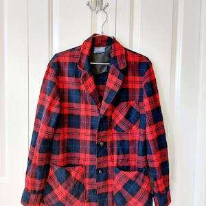 May include: A red and navy plaid blazer with two front pockets and a chest pocket. The blazer has a notched collar and three buttons. The jacket is hanging on a silver hanger on a white door.
