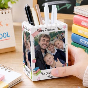 May include: A white square pen holder with a brick-like top edge, featuring a printed photograph of a teacher and students. The text "Happy Teachers' Day!" and "The Best Teacher Victor" are printed on the sides, decorated with small colorful illustrations of books, apples, and paperclips.