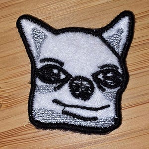May include: Embroidered patch featuring a stylized white dog face with black details. The dog has large eyes, a black nose, and a smiling mouth. The patch has a black border and is likely for clothing or accessories.