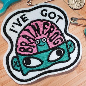 Könnte beinhalten: Gestickter Aufnäher mit dem Text "I'VE GOT BRAINS" mit einem Cartoon-Gehirn- und Gesichtsdesign. Das Gehirn ist rosa, das Gesicht ist türkis und der Aufnäher hat einen weißen Rand mit schwarzen Nähten. Der Aufnäher liegt auf einer Holzoberfläche mit Nähutensilien.