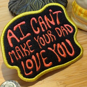 May include: A black embroidered patch with a yellow border featuring the orange text AI CAN'T MAKE YOUR DAD LOVE YOU. The patch is positioned next to a United States quarter coin for scale, showing the relative size of the embroidered fabric item.