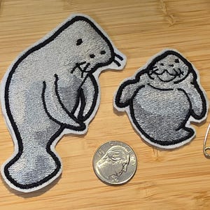 May include: Two embroidered patches featuring gray manatees with black outlines. The larger patch shows a side profile of a manatee, while the smaller patch shows a front-facing view. A silver US quarter coin is placed between them for scale.
