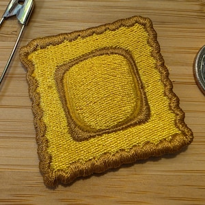 May include: A square yellow embroidered patch with a raised center and scalloped edges, positioned next to a silver safety pin and a US quarter coin for scale, showcasing detailed golden thread work and a textured, dimensional appearance.