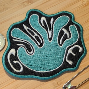 May include: An embroidered patch featuring a black background with a teal, hand-shaped design outlined in white thread. The patch has a teal border and is displayed next to a silver coin and a metal safety pin for scale on a wooden surface.
