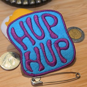 May include: A light blue embroidered patch with a purple border and the purple text HUP HUP. The patch is positioned next to a silver-colored quarter coin, a gold-colored coin, and a silver metal safety pin on a light wood surface.