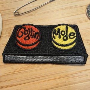 May include: A rectangular black embroidered patch featuring two circular motifs. The left circle is orange with the text "Gollin" in black, and the right circle is yellow with the text "Moje" in black. A grey horizontal line runs along the bottom.