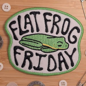 May include: An oval-shaped embroidered patch with a white background and a green border. The patch features a green frog illustration in the centre with the black embroidered text "FLAT FROG FRIDAY" arched above and below the frog graphic.